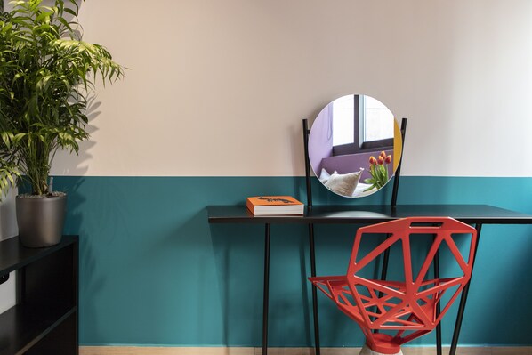 Una habitación azul verdoso y blanca con un escritorio negro, una silla roja, una planta en maceta y un espejo redondo que refleja una ventana con una planta.