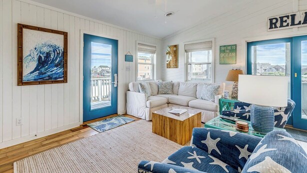 A coastal-themed living room with a white and blue color scheme, featuring a white sofa, a blue sofa with star patterns, a wooden coffee table, and a ceiling fan. - Blue Bungalow | 625 ft from the beach | Hot Tub, Dog Friendly | Kitty Hawk (Kitty Hawk)