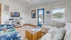 A spacious living room with a white wooden wall, a ceiling fan, a large window, a beige sofa, a coffee table, and a TV on a wooden stand. - Blue Bungalow | 625 ft from the beach | Hot Tub, Dog Friendly | Kitty Hawk (Kitty Hawk)