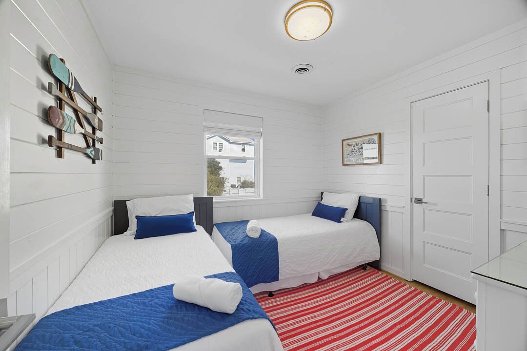 A room with two beds, white walls, a window, a door, and a red-striped rug.