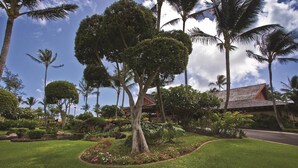 Um jardim tropical com gramado bem cuidado, diversas árvores e um edifício com telhado vermelho.