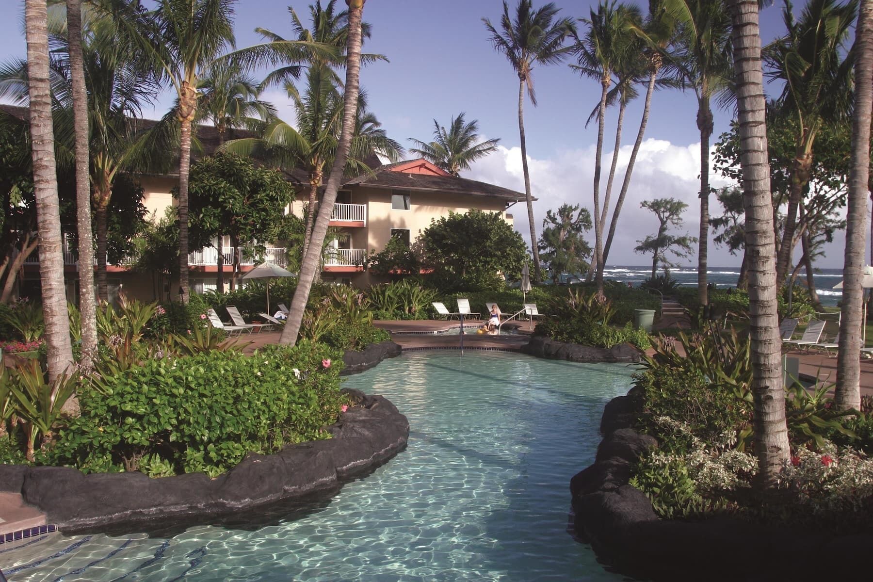 A pool surrounded by palm trees and a building with a red roof.