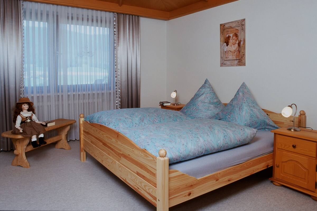 Een houten bed met een blauw beddegoed met patroon, een nachtkastje met een lamp, een houten bankje en een raam met dunne gordijnen.