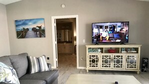 Ein Wohnzimmer mit grauem Sofa, einem weißen Couchtisch mit Meerfunde-Dekoration, einem Fernseher auf einer Kommode und einem Gemälde mit Strandmotiv.