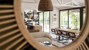Un espace de repas moderne avec une grande table et des chaises en bois, un luminaire suspendu en fibres tressées et de grandes fenêtres offrant une vue sur la verdure.