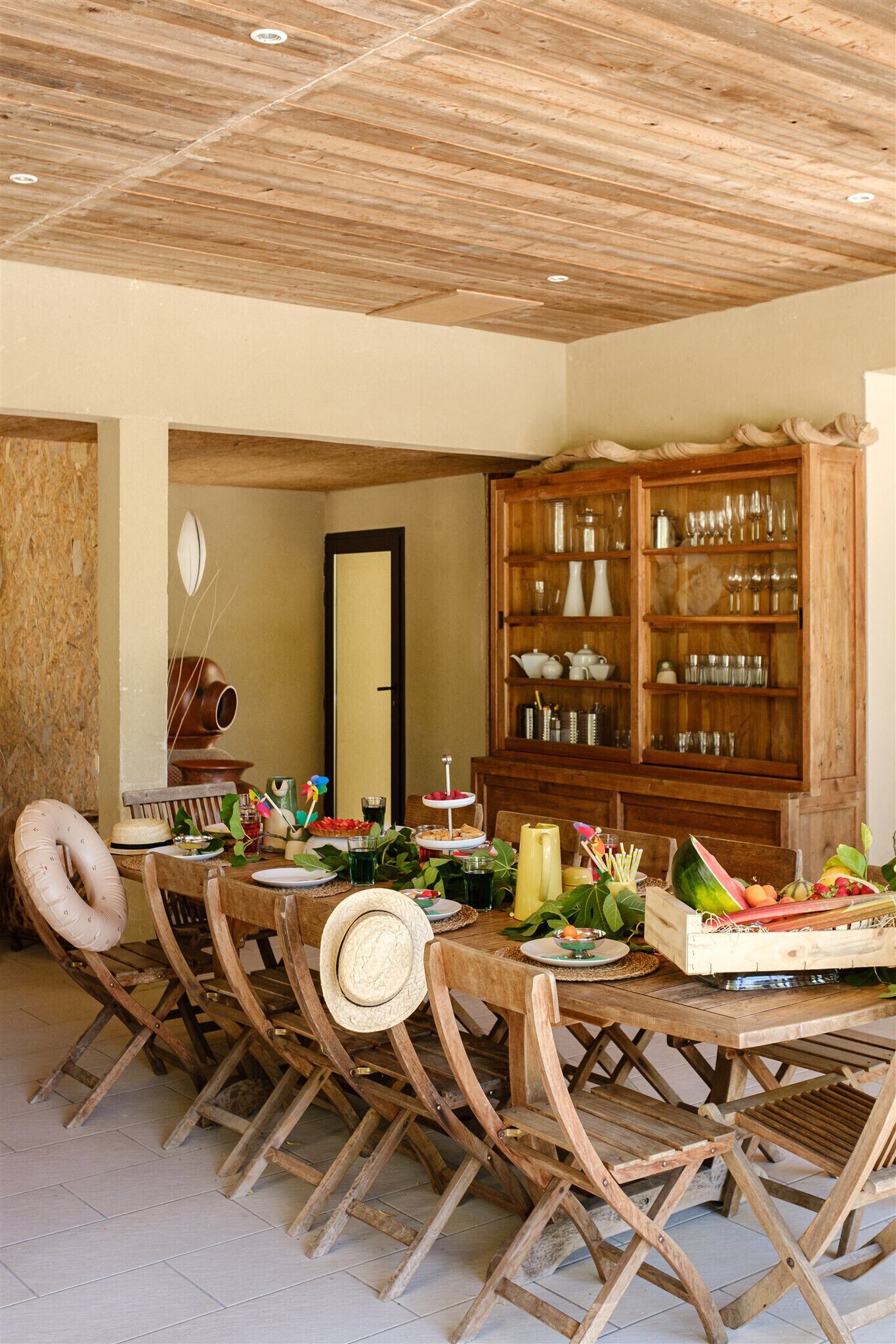 Un coin repas avec une table et des chaises en bois, un buffet en bois contenant de la verrerie, et un plafond rustique.