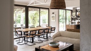 Un espace repas avec une table et des chaises en bois, un luminaire suspendu en fibres tressées et une vue sur l’extérieur grâce à de grandes fenêtres.