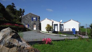 Une maison en pierre à deux étages, avec une allée en gravier, entourée de verdure et de fleurs.