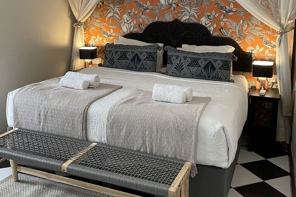 A bed with a canopy, patterned pillows, a bedside table with a lamp, and a bench.