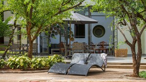 Un área junto a la piscina con sillones de descanso, una mesa de comedor y sillas, rodeada de vegetación y un edificio con placas decorativas en la pared.
