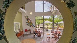 Vista circular de un comedor con mesas y sillas, una pared con platos de colores, y un amplio ventanal con vistas a un jardín.