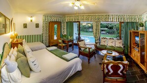 A hotel room with a bed, a seating area with chairs and a table, a wooden cabinet, and a view of the garden through the windows. - Wilmot Cottages (Gqeberha (Port Elizabeth))