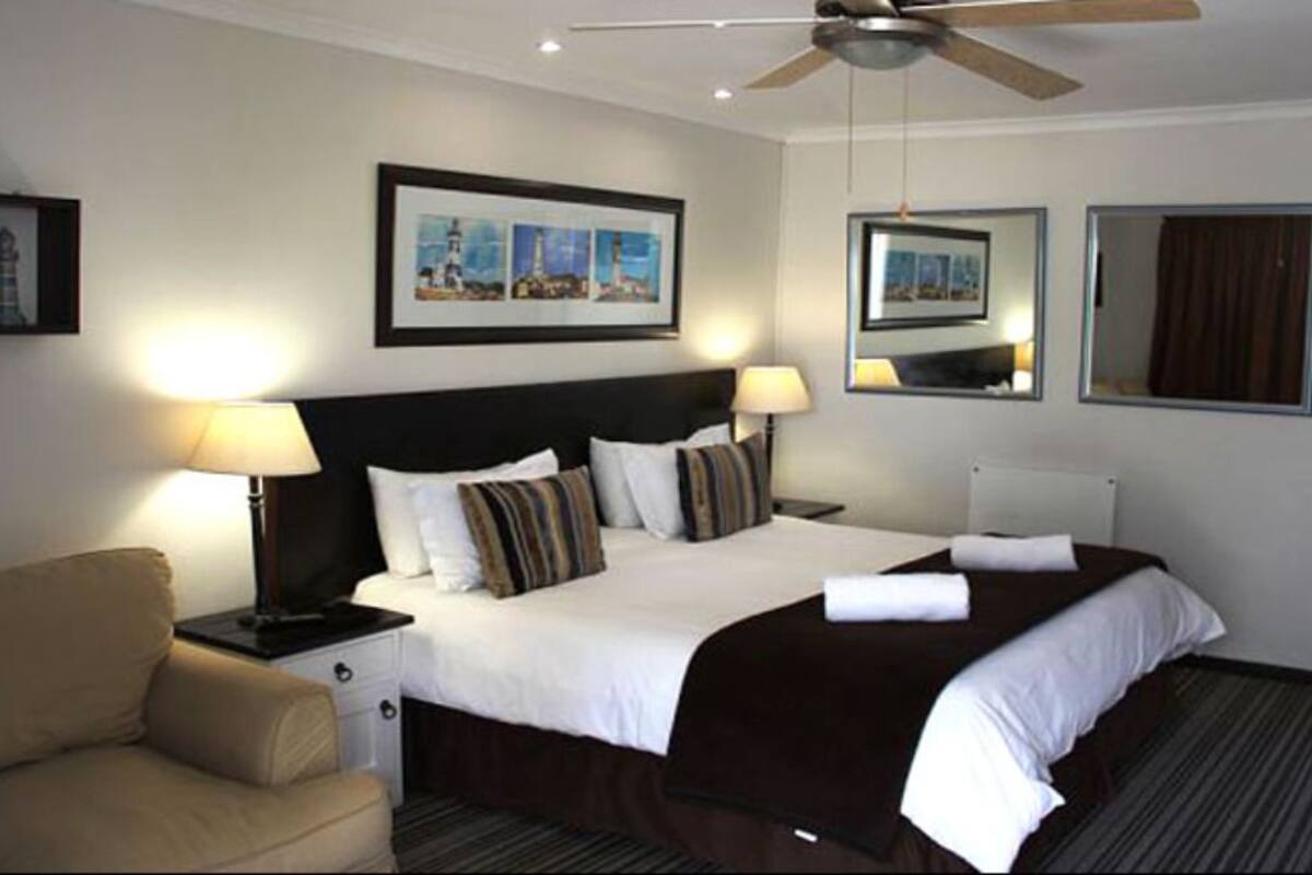 Una habitación de hotel con una cama grande, dos lámparas de noche, un ventilador de techo e imágenes enmarcadas en las paredes.