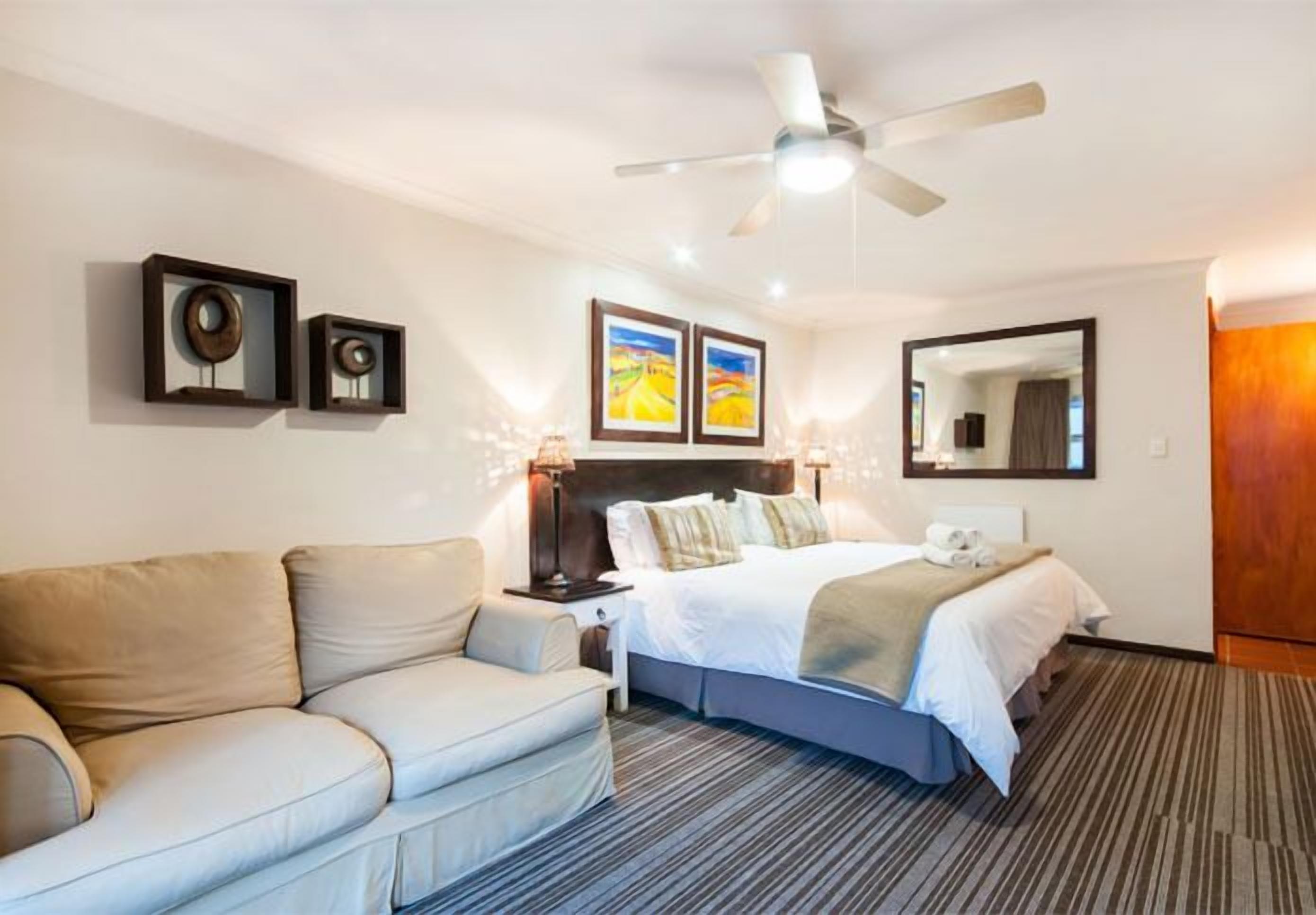 A hotel room with a bed, a sofa, a ceiling fan, framed artwork, and a mirror.