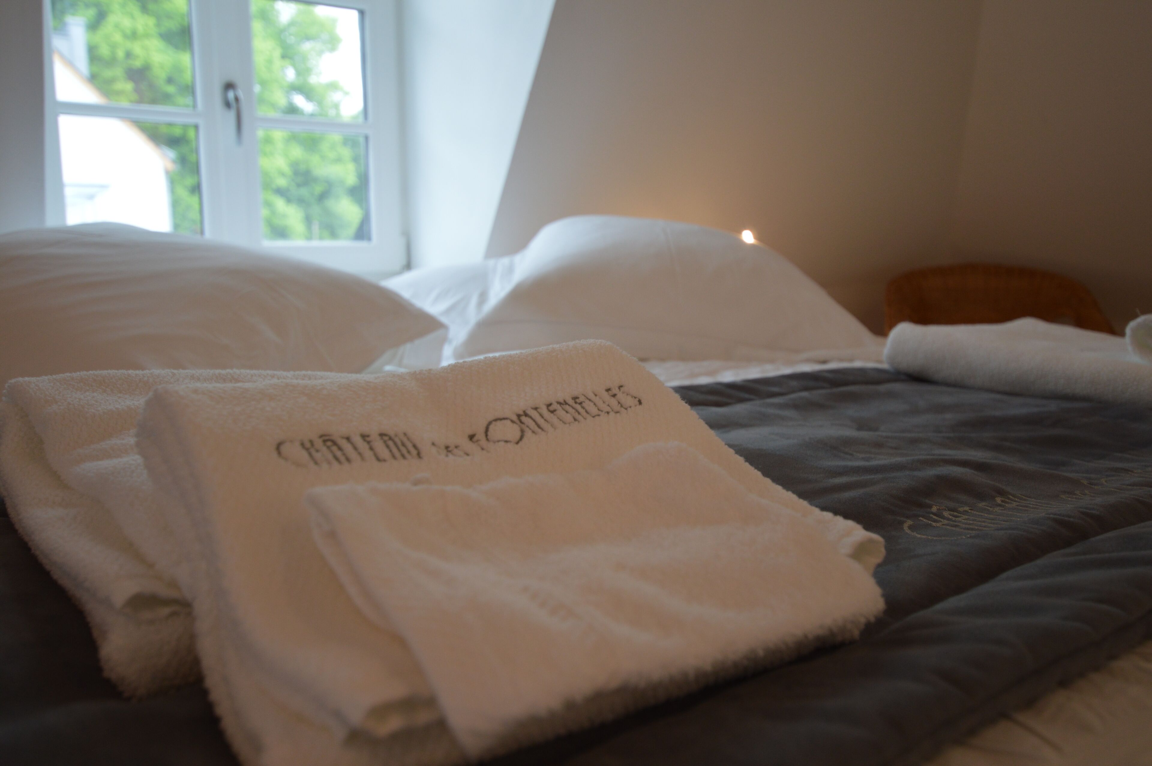 A neatly made bed with white linens and a folded towel with text, a window with a view of trees, and a bedside table with a lamp.