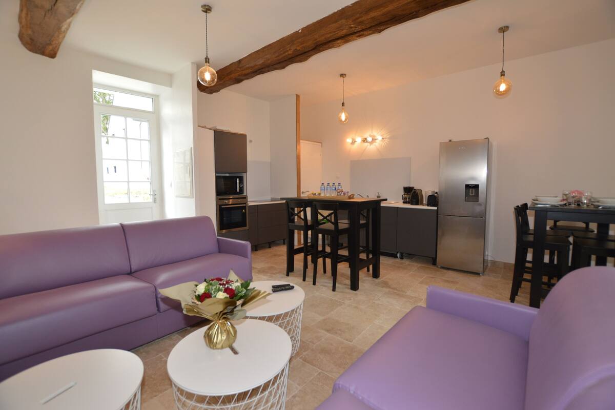 A modern living room with a purple sofa, a dining table, and a kitchen area with a refrigerator and microwave.