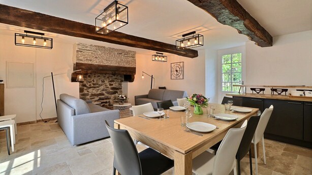 Un comedor con una mesa de madera dispuesta para cuatro personas, rodeada de sillas. Se ve una chimenea de piedra y un sofá al fondo.
