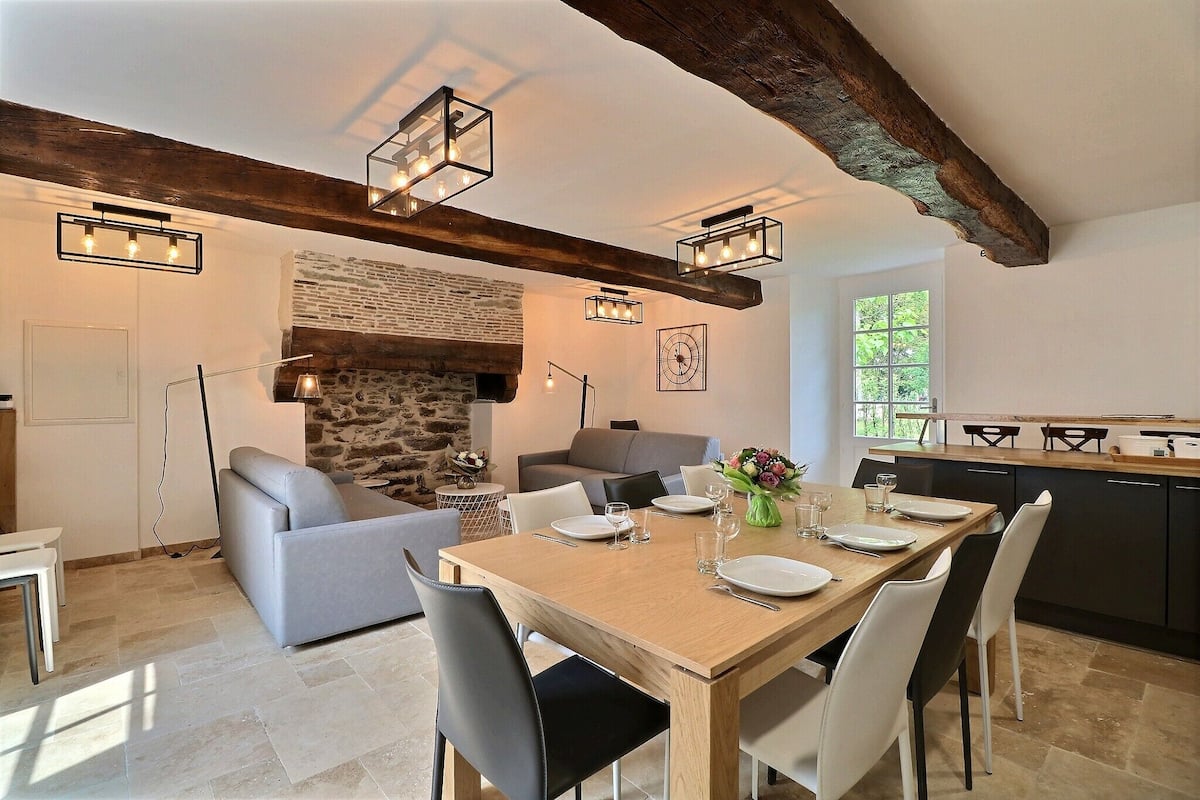 A dining area with a wooden table set for four, surrounded by chairs. A stone fireplace and a sofa are visible in the background.