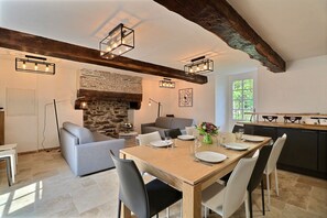 Un comedor con una mesa de madera dispuesta para cuatro personas, rodeada de sillas. Se ve una chimenea de piedra y un sofá al fondo.