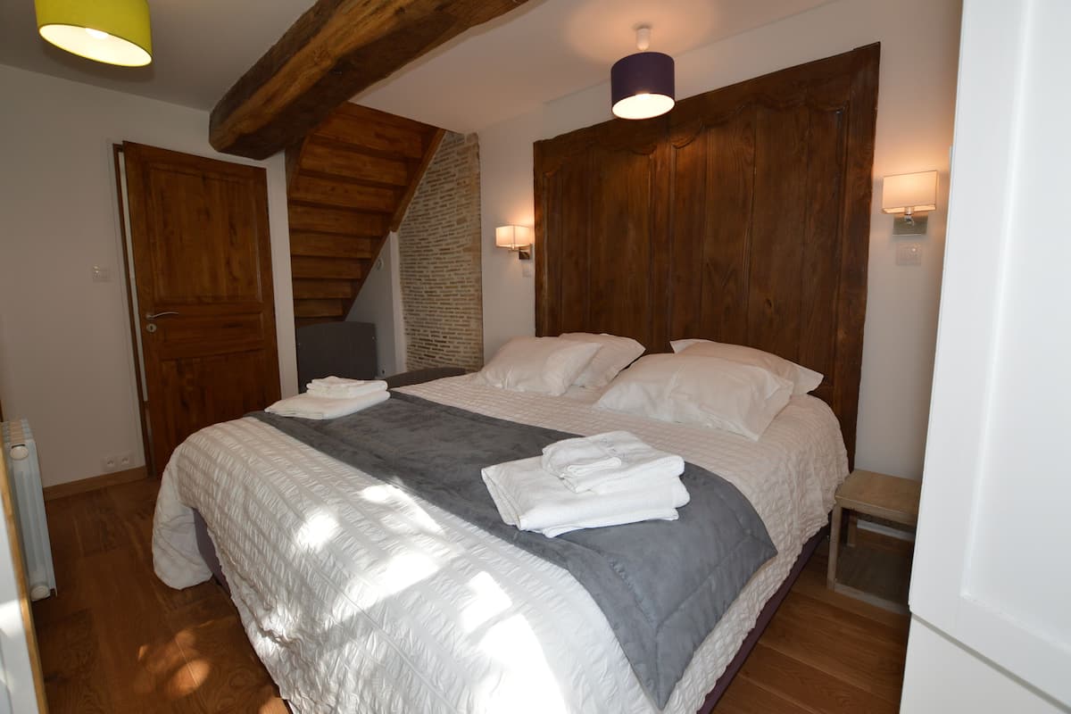 A bedroom with a large bed, wooden headboard, a small wooden stool, and a staircase.