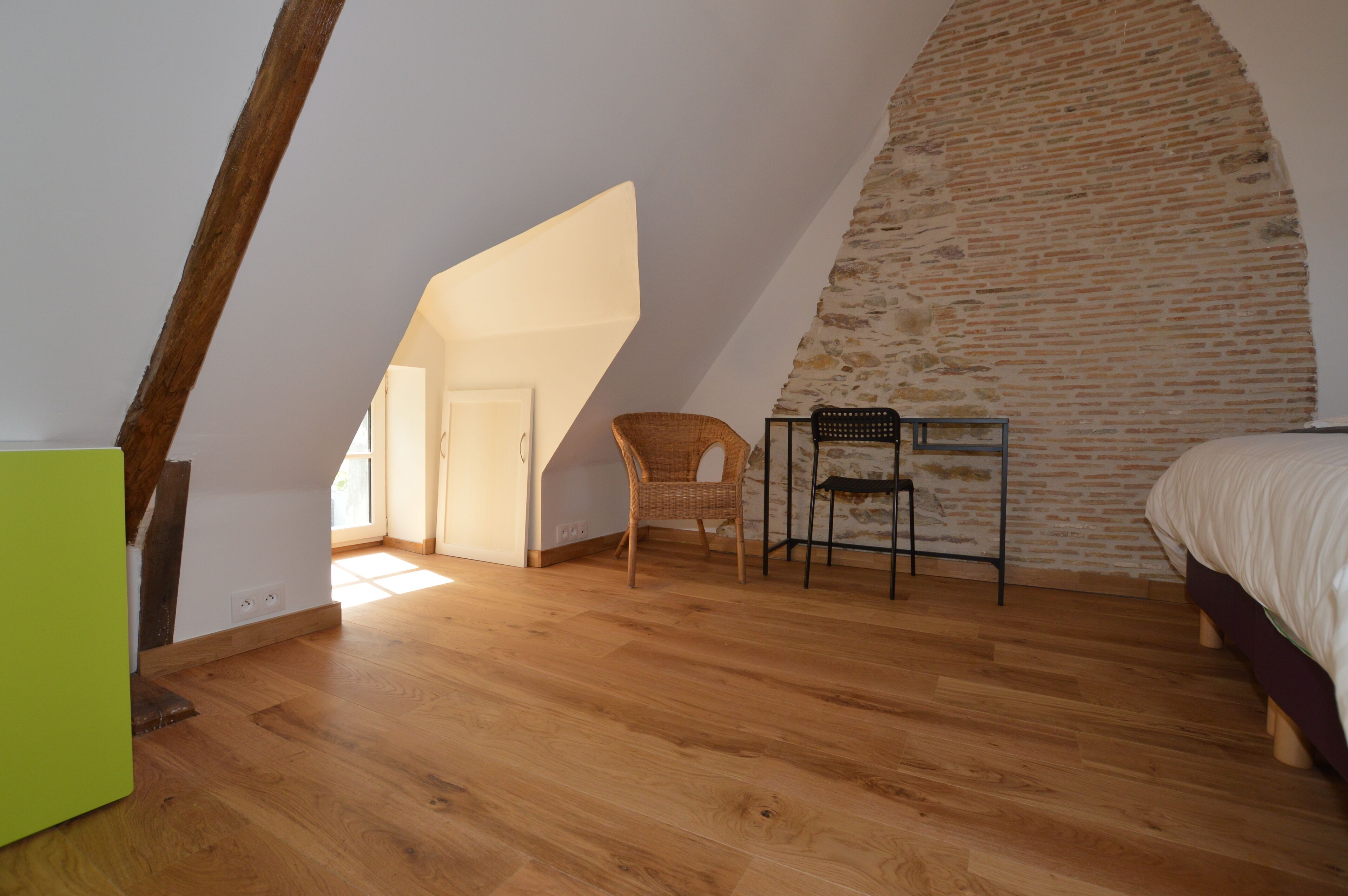 Un dormitorio con techo inclinado, suelo de madera, cama, silla y una mesita.