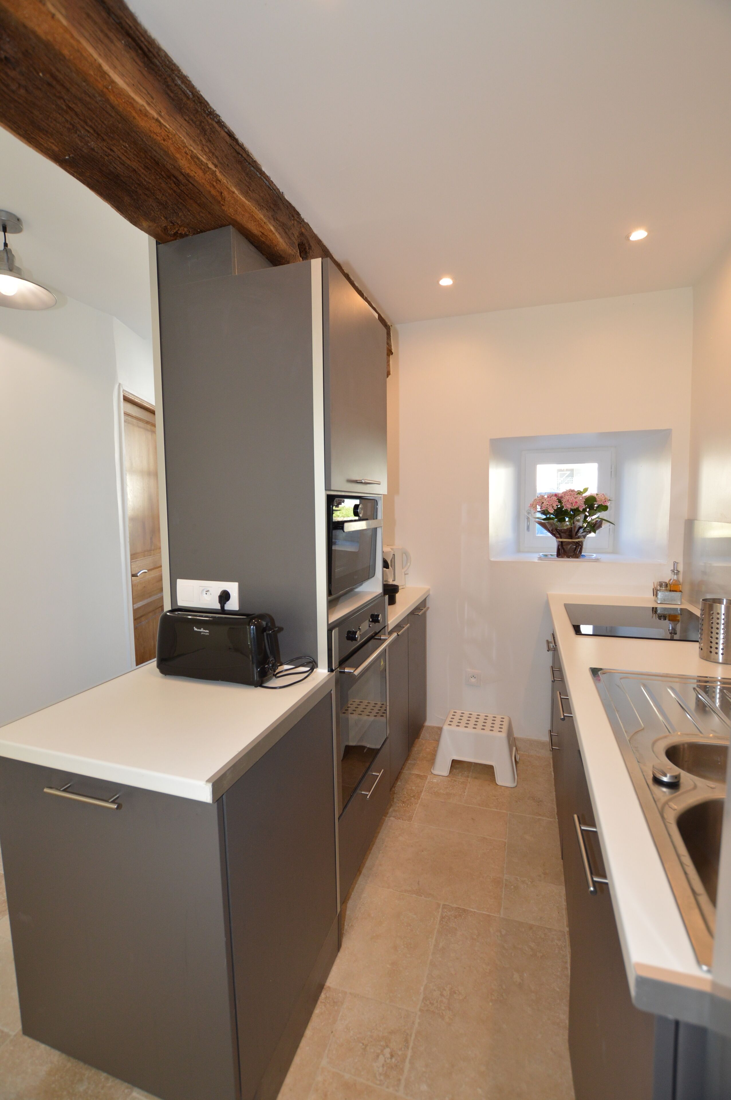 A compact kitchen with modern appliances, a small window with a flower vase, and a wooden beam ceiling.