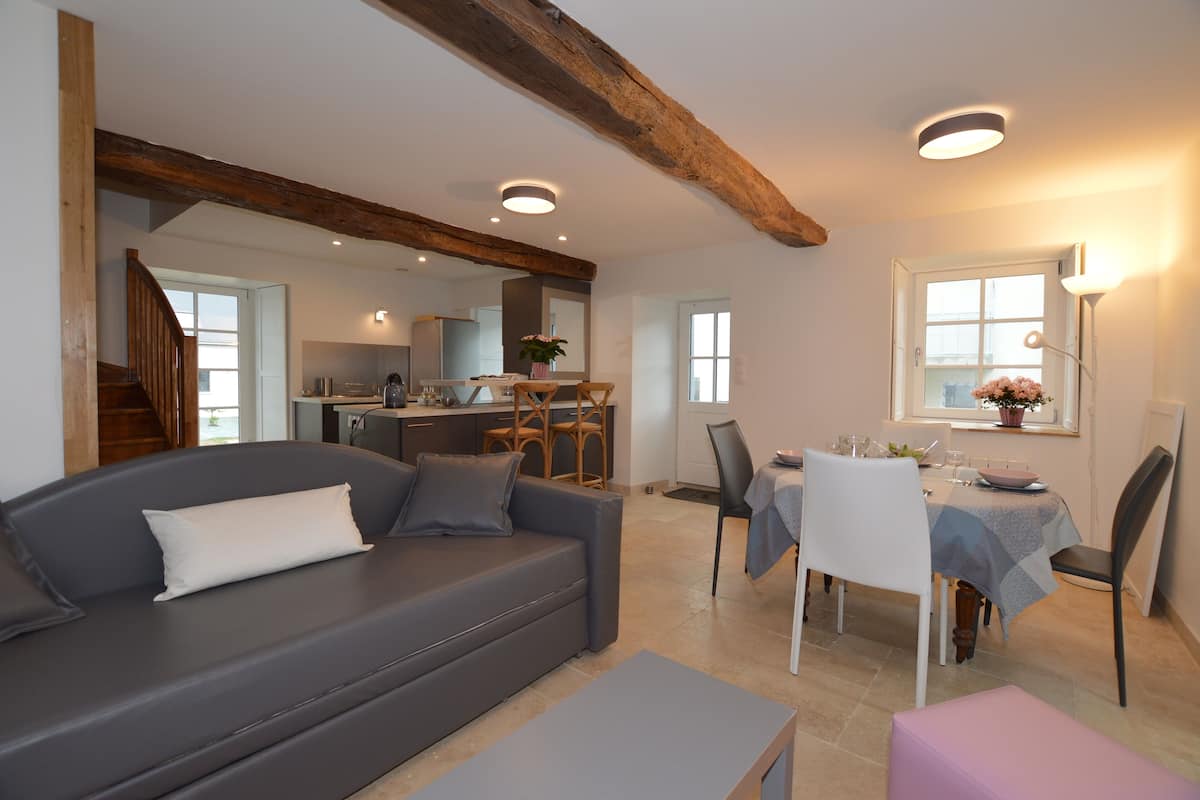 A modern living room with a grey sofa, wooden beams, a dining area with a table and chairs, and a kitchen in the background.