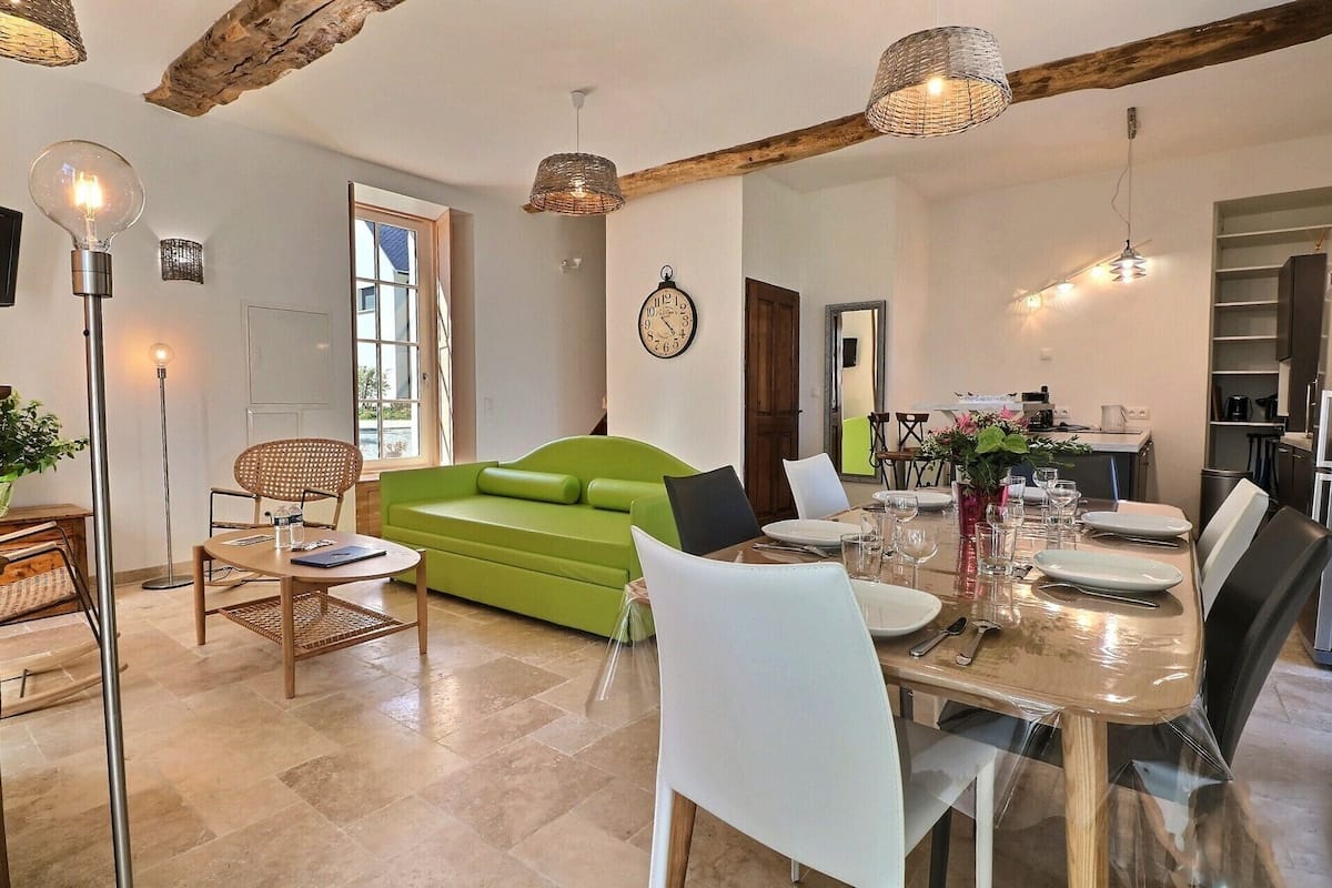 A modern living room with a green sofa, dining table set for six, and a kitchen in the background.