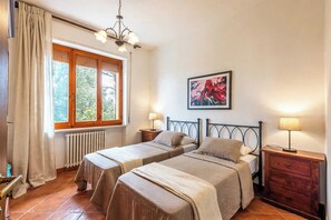 Een slaapkamer met twee bedden, een radiator, een raam met gordijnen, een kroonluchter, een nachtkastje met een lamp en een schilderij aan de muur.