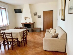 Een woonkamer met een open haard, een bank, een eettafel met stoelen en een aan de muur bevestigde televisie.