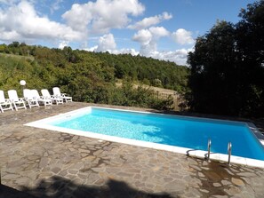 Une piscine rectangulaire avec une terrasse en pierre, entourée de chaises longues blanches et d’une clôture en métal.