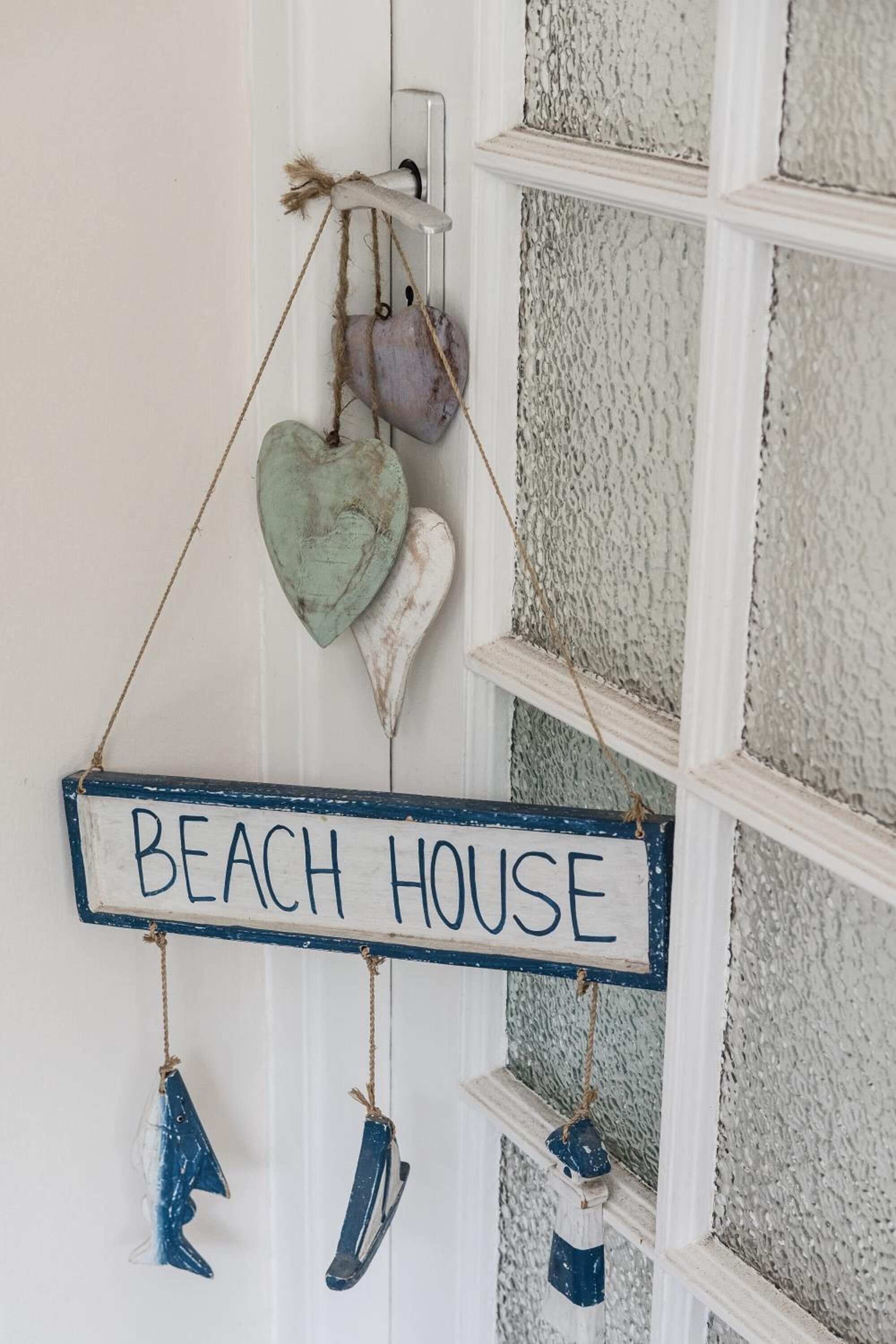 A sign with the words "BEACH HOUSE" hanging on a white door with a textured wall.