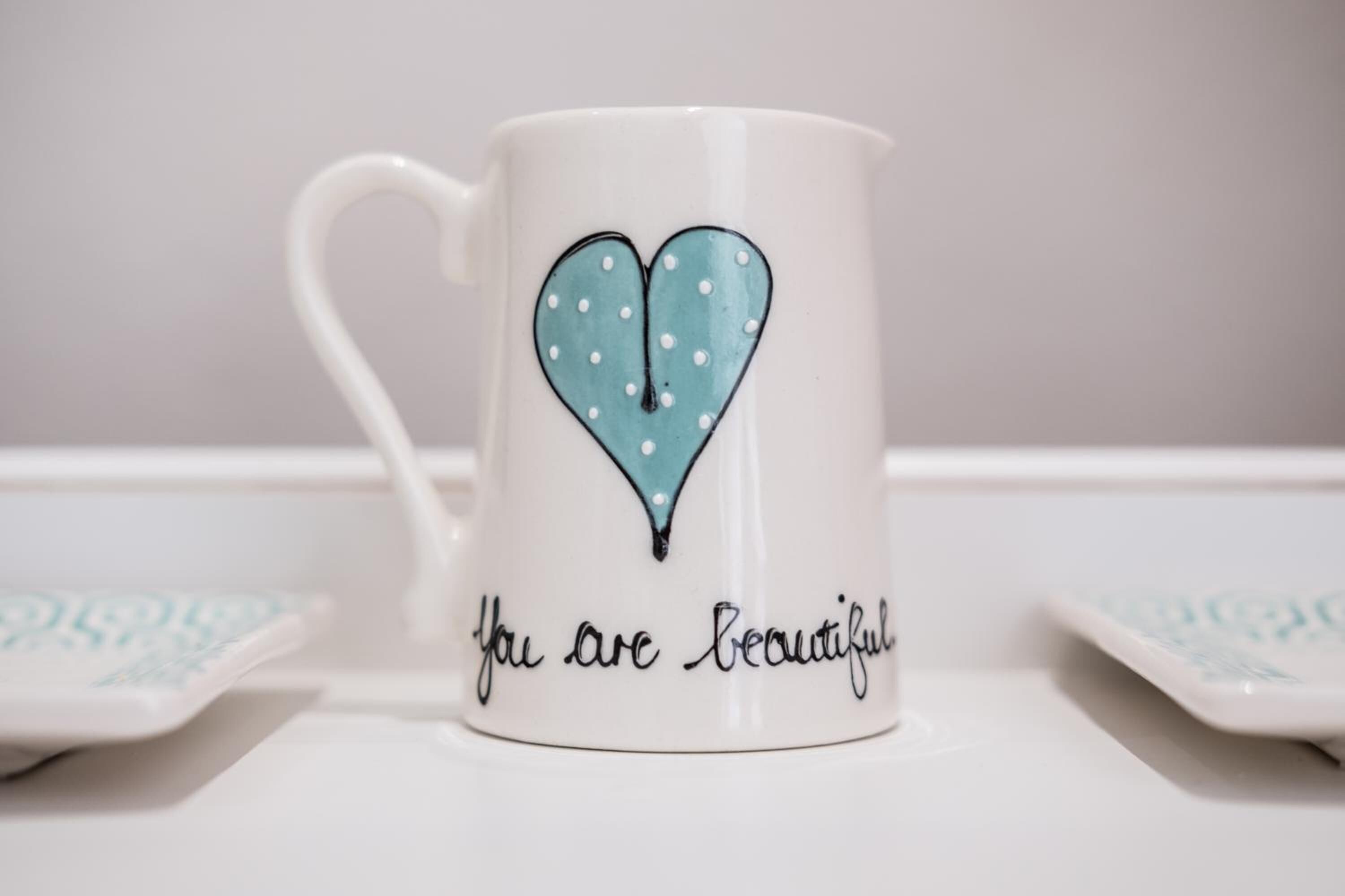 A white ceramic mug with a heart design and the text "you are beautiful" on a white surface.