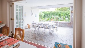 An open-plan living space with a dining area, a kitchen, and a patio with a view of a lush garden. - On The Bay B&B (Gqeberha (Port Elizabeth))