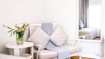 Una habitación con un sofá blanco, una mesa redonda con un jarrón de flores rosadas y un espejo que refleja una cama y cortinas.