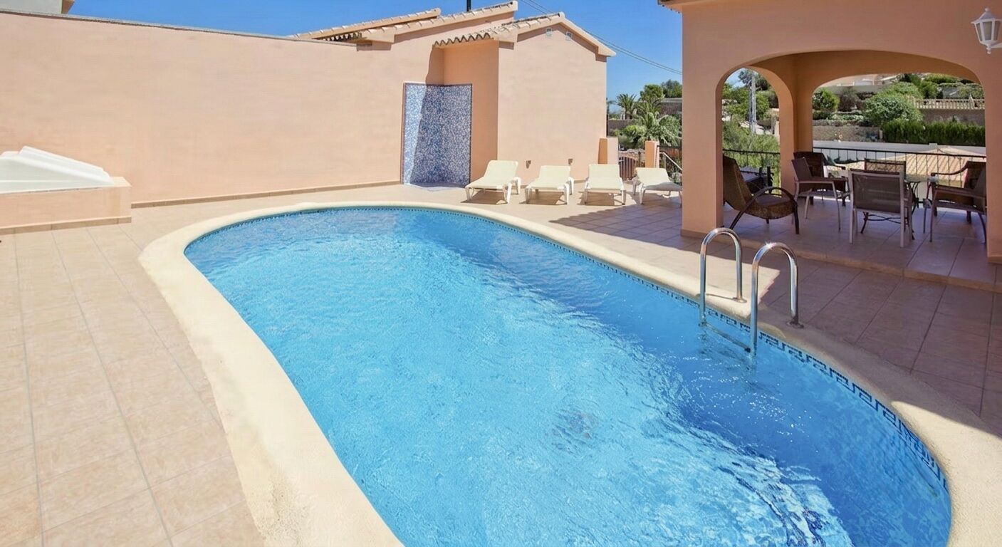 A swimming pool with a clear blue water, surrounded by a beige tiled deck, and a patio area with chairs and a table.