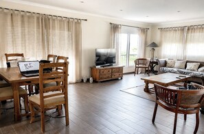 Een woonkamer met een houten eettafel, stoelen, een bank, een televisie en een lamp.