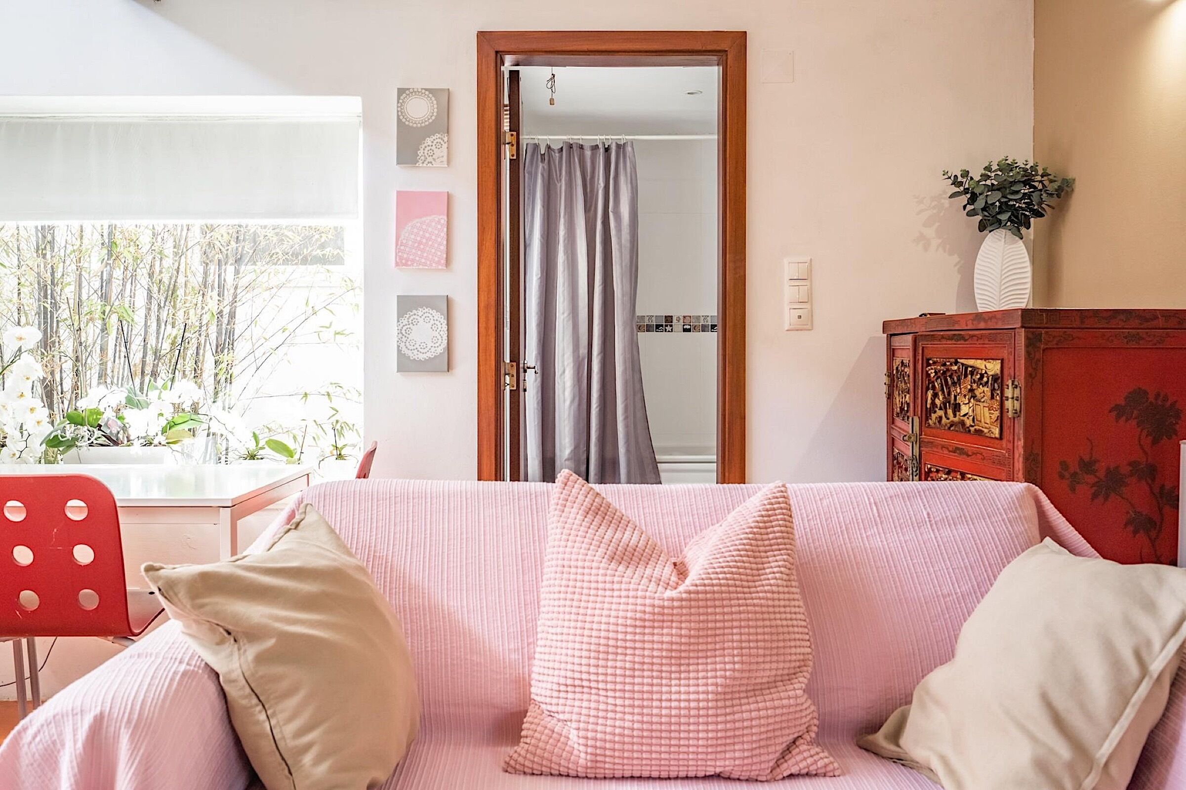 Une pièce avec un canapé rose, un fauteuil rouge, un meuble en bois et une vue sur un jardin de bambous, visible par une fenêtre.