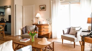 Un salon chaleureux avec un canapé, deux fauteuils, une table basse et une petite table en bois. On y trouve une lampe, un tableau au mur et une fenêtre avec des rideaux.