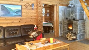 A cozy log cabin interior with a fireplace, wooden walls, a leather sofa, and a dining table with a chessboard and fruit. - Villa Luppopirtti (Sodankyla)