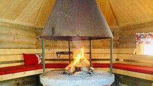 A wooden cabin interior with a central fire pit, benches, and a wooden ceiling. - Villa Luppopirtti (Sodankyla)