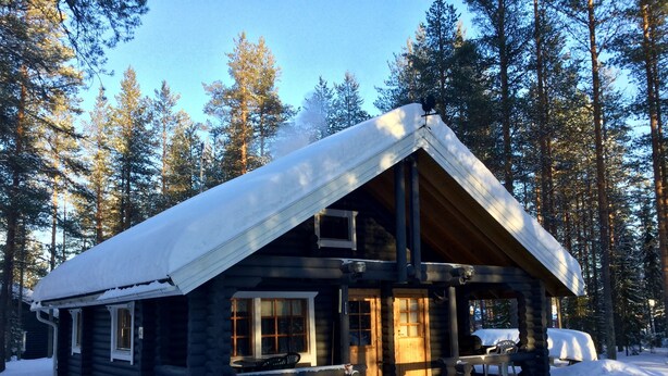 A log cabin with a snow-covered roof, a porch with a bench, and a window overlooking a forest. - Villa Luppopirtti (Sodankyla)