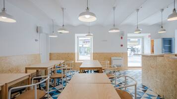 A spacious cafe with multiple wooden tables and chairs, white pendant lights, and a geometric patterned floor.