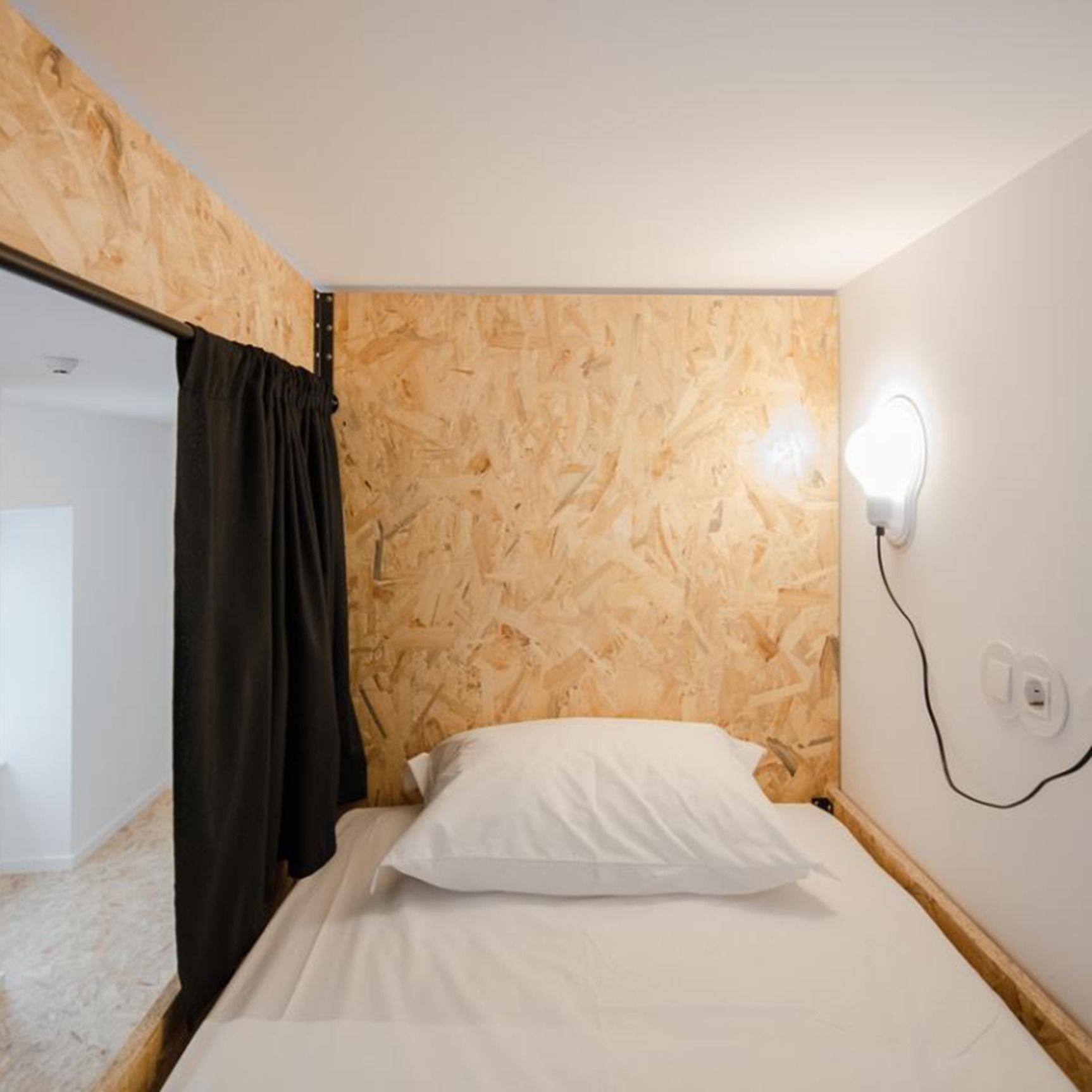 A bedroom with a wooden wall, a bed with white bedding, a black curtain, and a wall-mounted light.