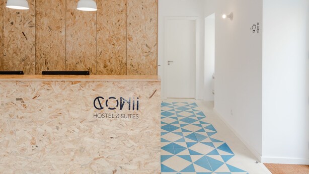 Un área de recepción con un mostrador de madera identificado como "COVII HOSTEL & SUITES", suelo con un diseño geométrico e iluminación colgante.