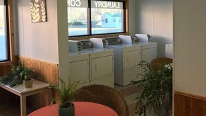 A room with a dining table, chairs, a kitchen area with appliances, and a window with a view of a house. - Wheels Motel (Greybull)