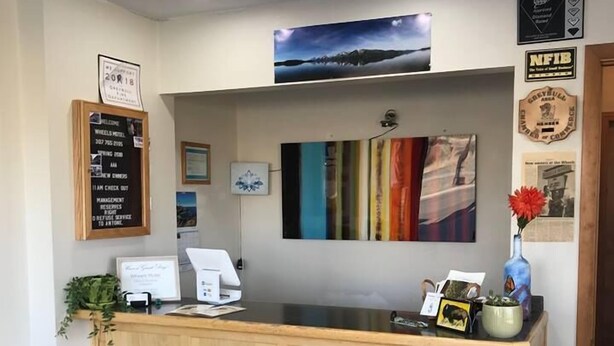 A reception desk with a wooden counter, a framed welcome sign, a framed picture, and a wall-mounted certificate. - Wheels Motel (Greybull)
