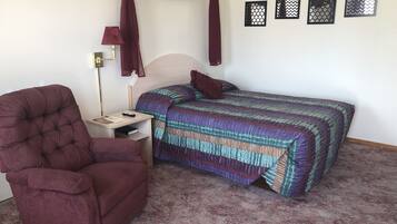 A bedroom with a tufted armchair, a bedside table with a lamp, a bed with a patterned bedspread, and four wall-mounted decorative items.