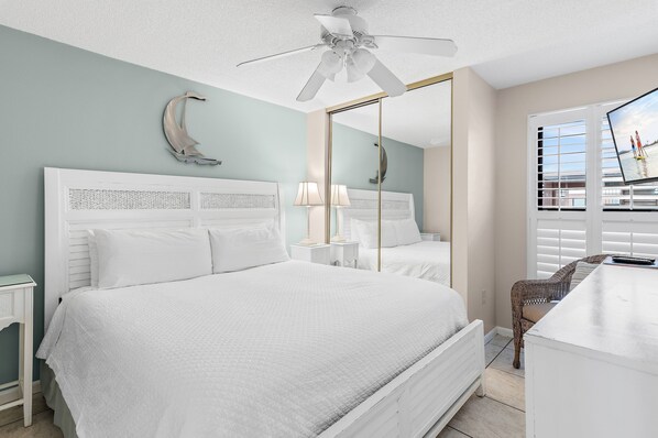 A bedroom with a bed, bedside tables, a ceiling fan, a large mirror, and a view of the outdoors through a window. - Open Concept floor plan, larger than a standard one bedroom! One free beach chai (Destin)