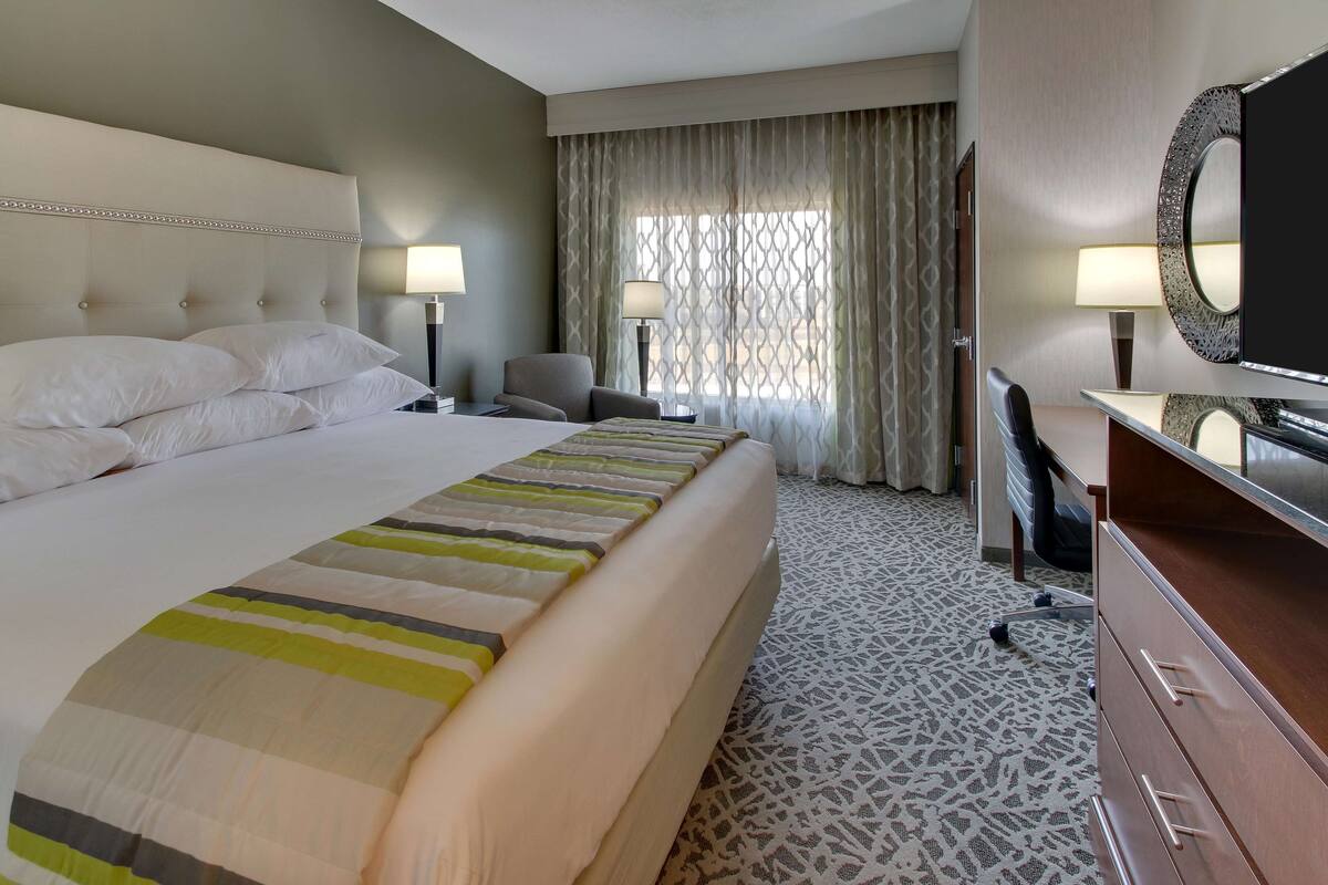 A hotel room with a large bed, two bedside tables with lamps, a desk with a chair, a TV, and a patterned carpet.